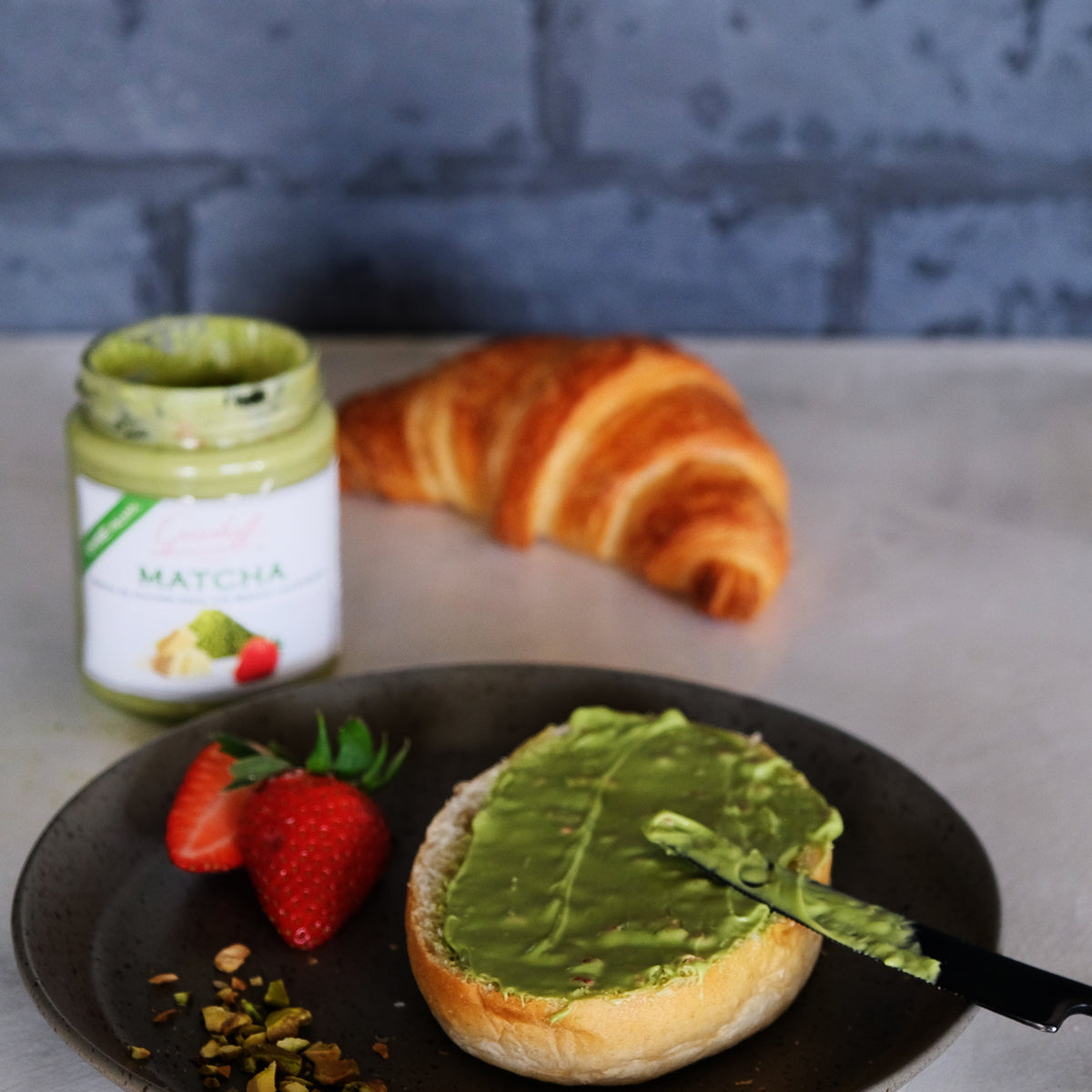 Ein Brioche bestrichen mit Matcha-Schokoladencreme. Im Hintergrund ein Croissant und ein Glas weiße Schokoladencreme mit Matcha und Erdbeeren von Grashoff.