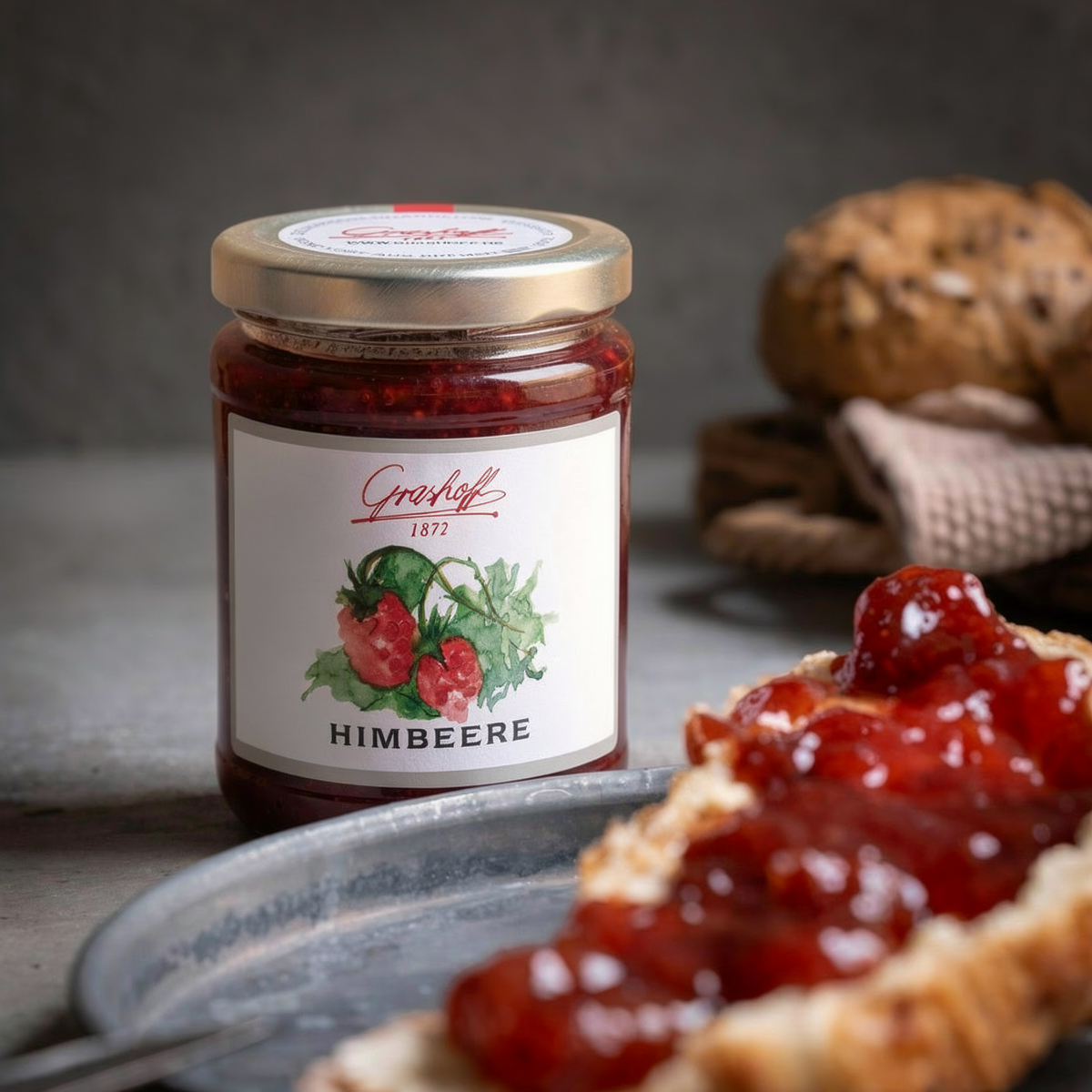 Ein Glas Himbeerkonfitüre von Grashoff neben einem Brötchen