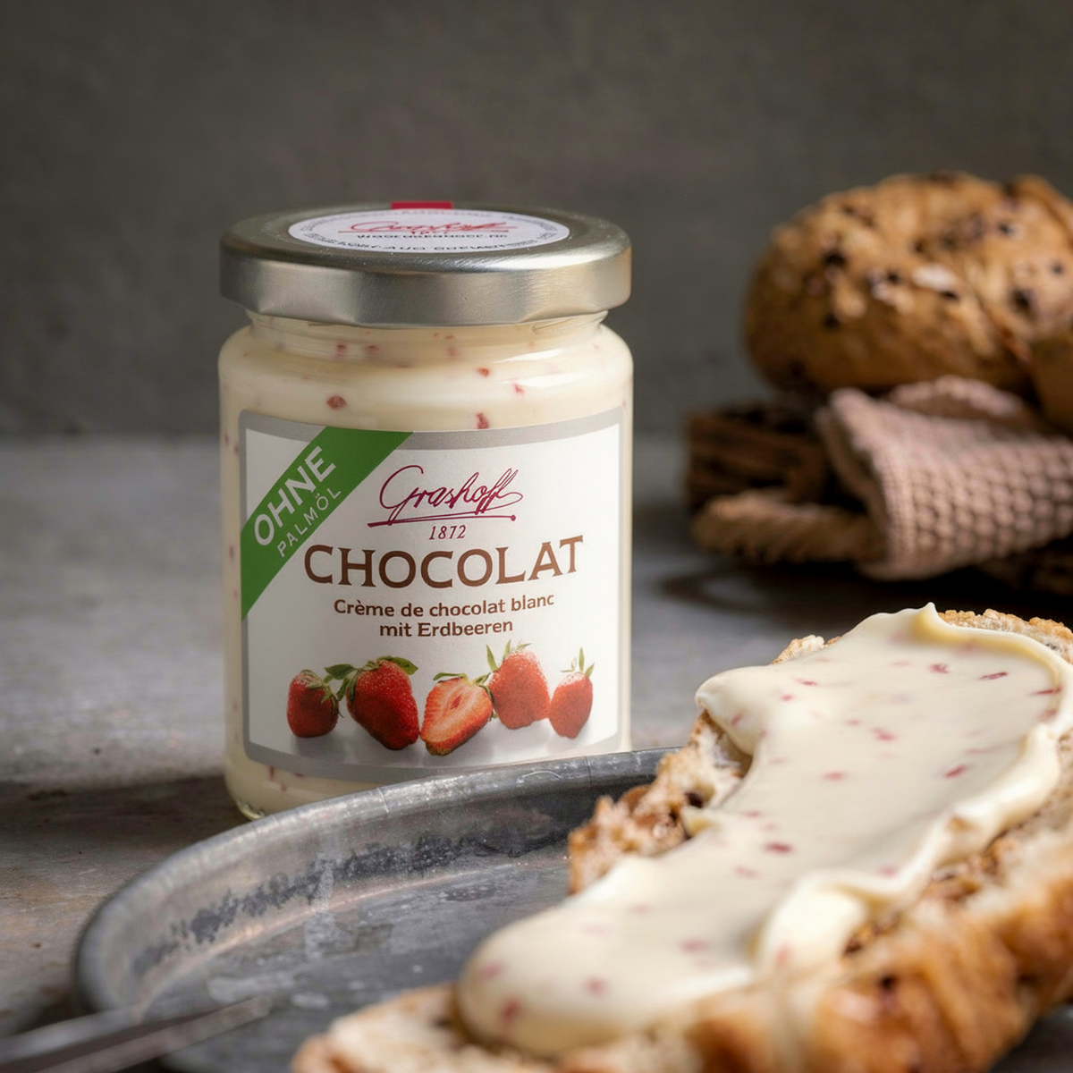 Ein Glas weiße Schokoladencreme mit Erdbeeren von Grashoff neben einem Brötchen.